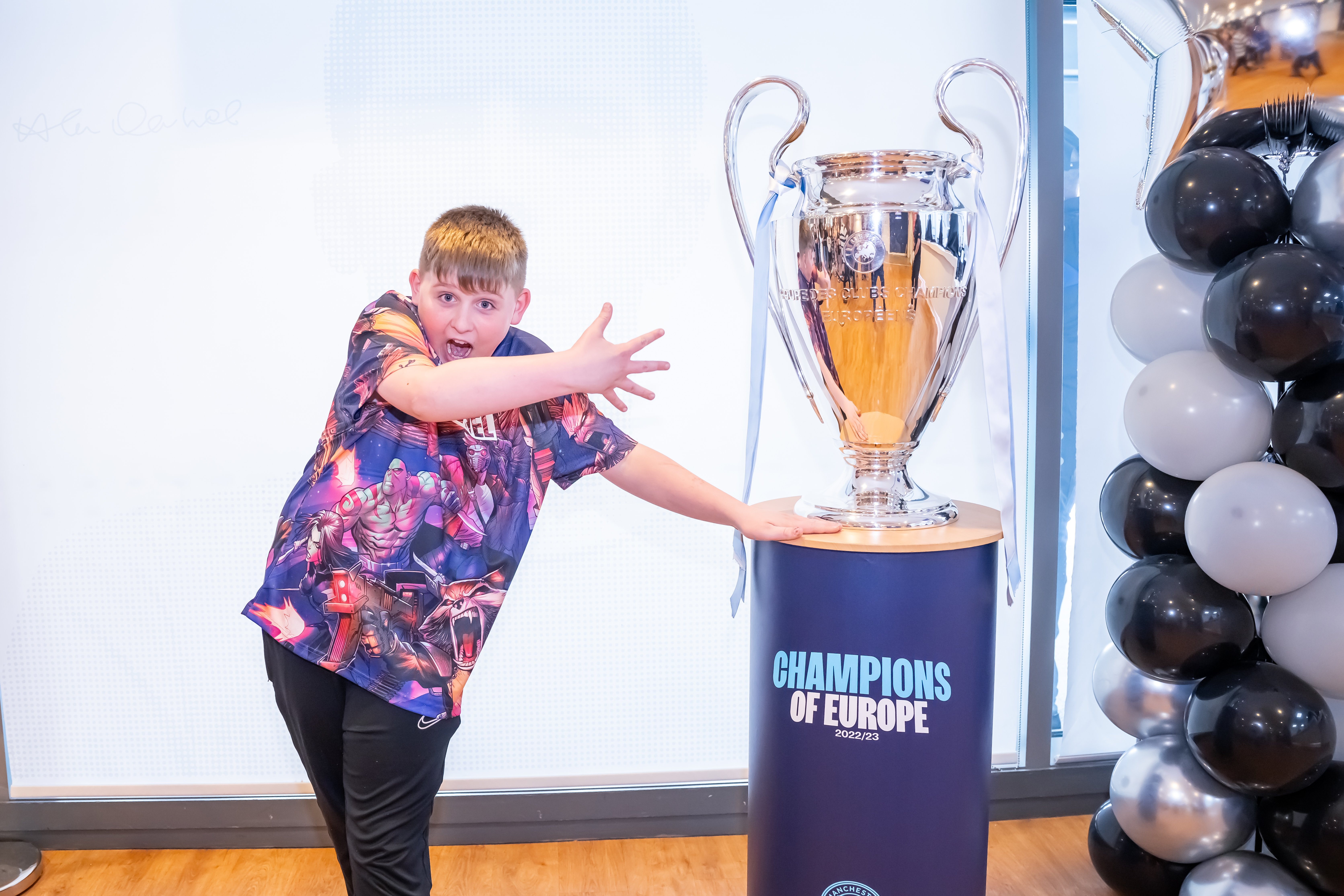 A young boy showing excitement and wide smile next to Manchester City’s Champions of Europe Cup 2022/23