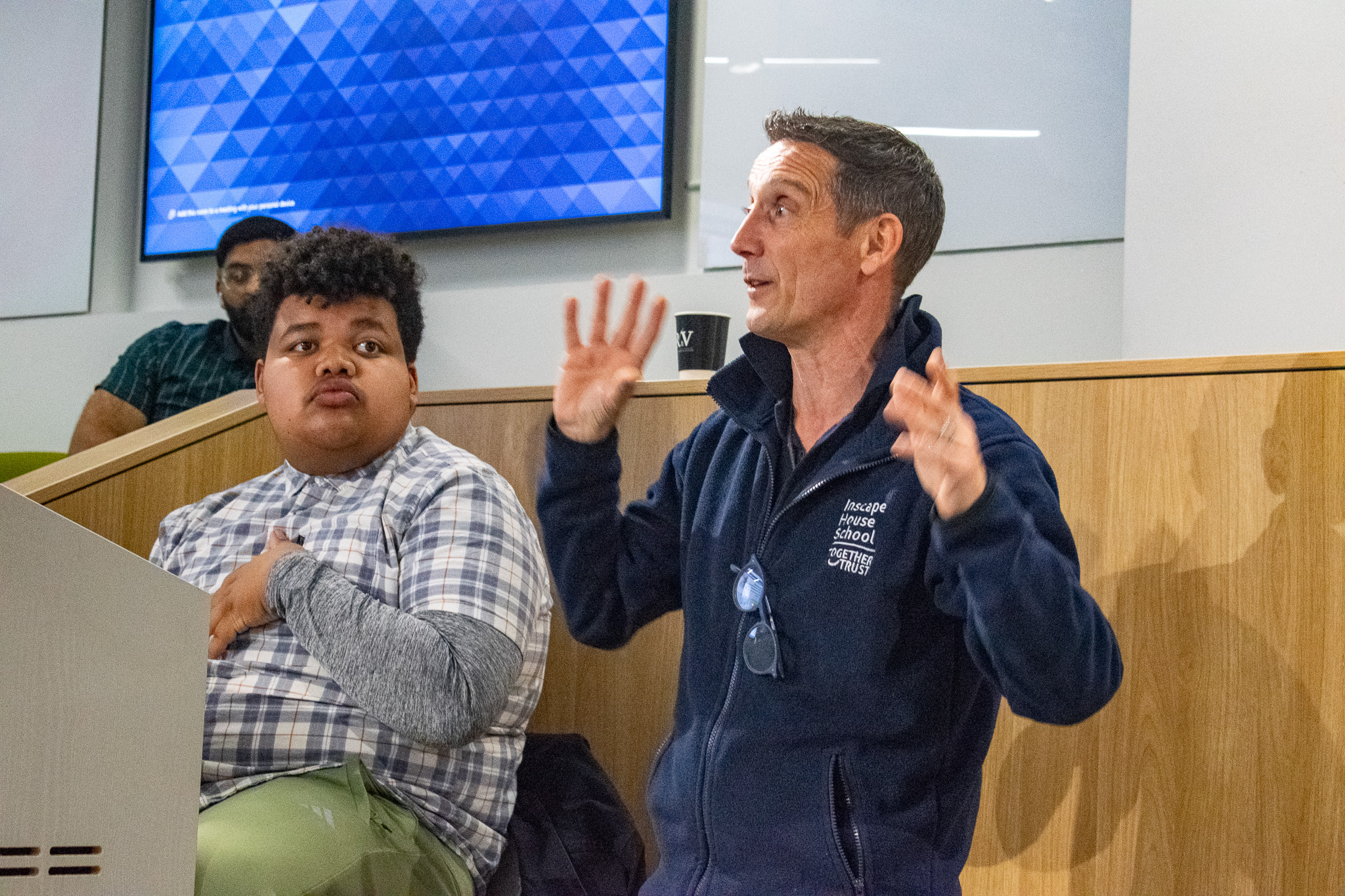 Greg speaking in front of a room, gesticulating, next to Mason who is operating a computer.