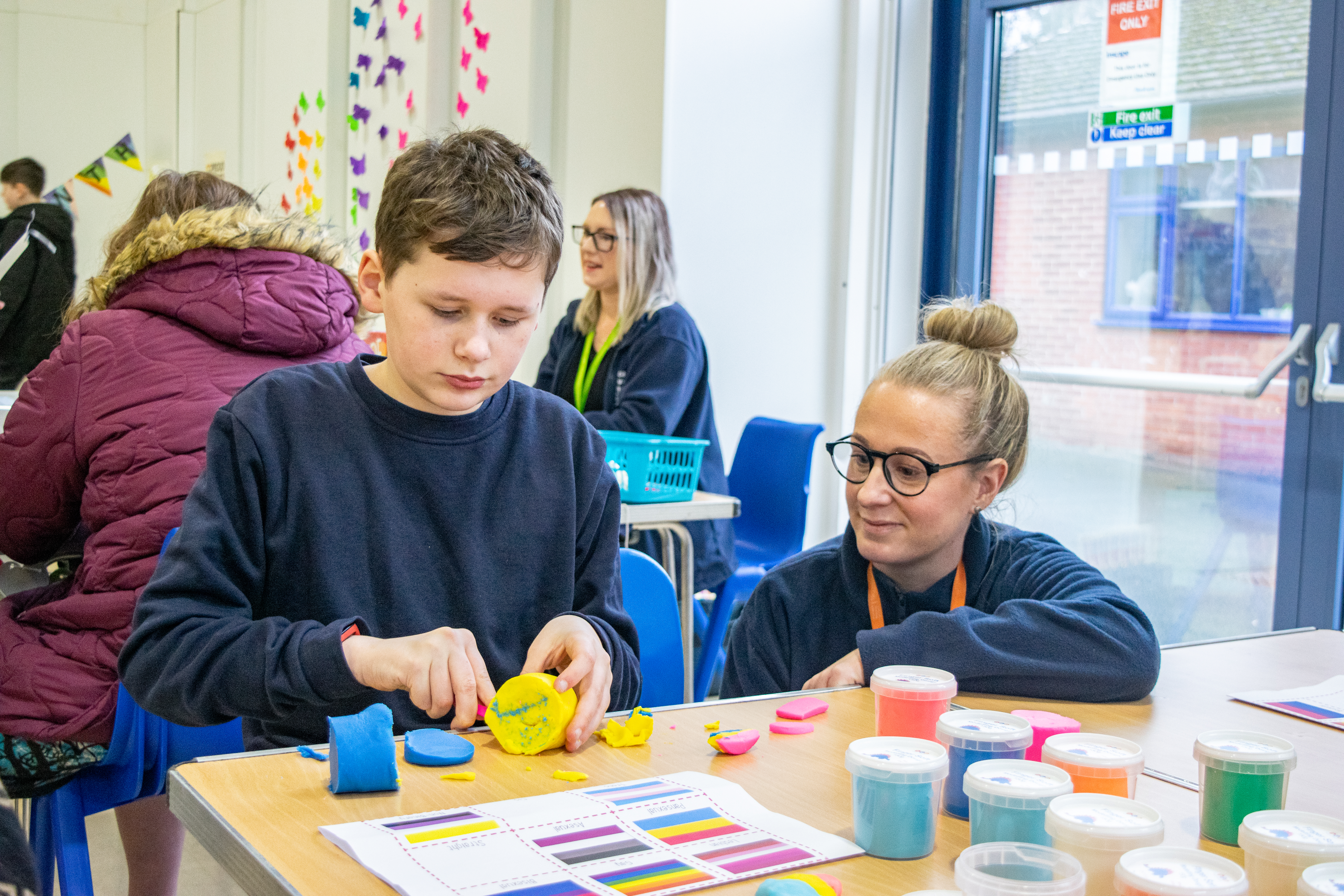 Young boy cutting into colorful play-doh clay. Next to him, a teacher is looking at his work smiling. 