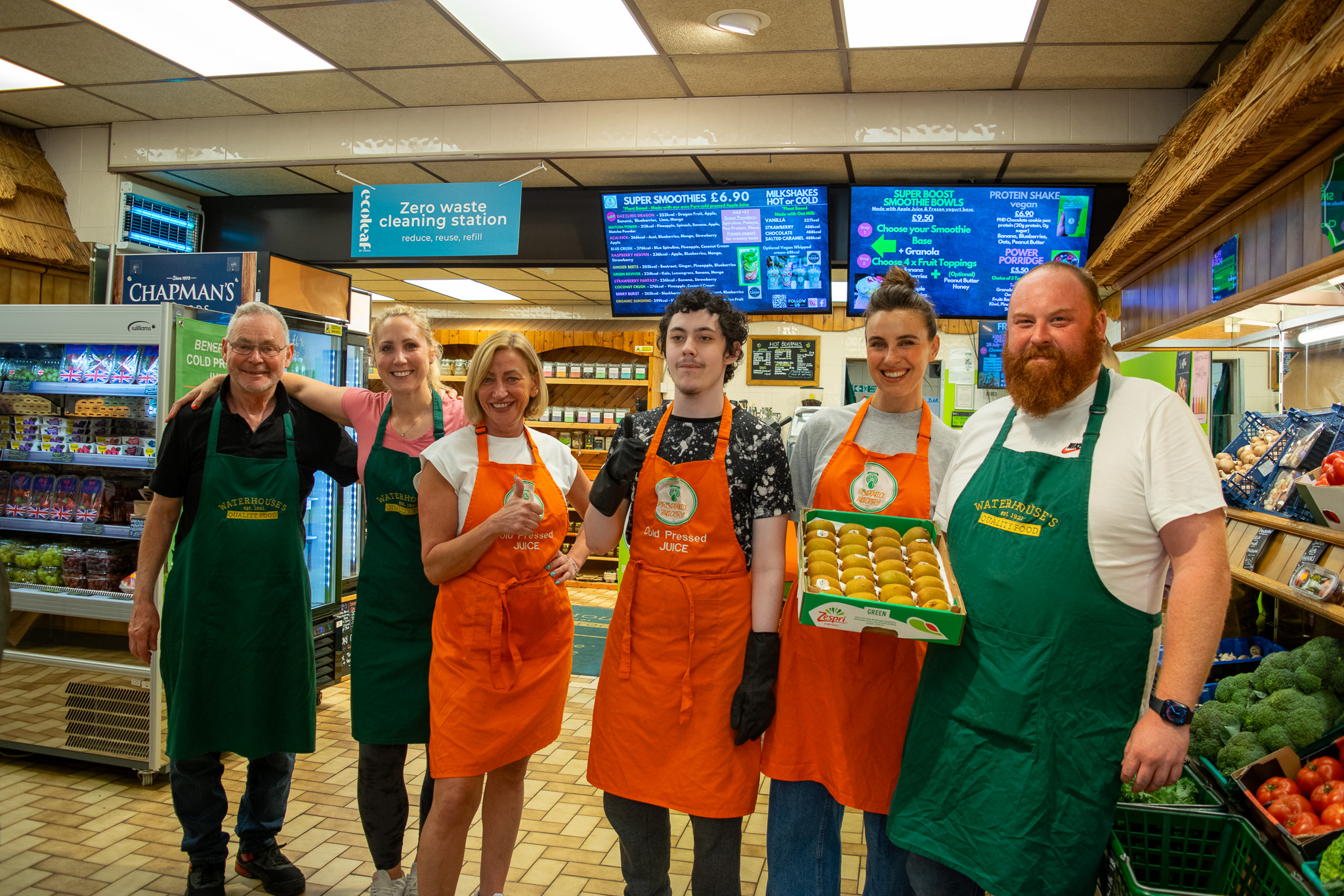 A teen boy is smiling next to 5 workers from the shop. They are posing inside the shop. There are fridges of fresh produce behind them. 