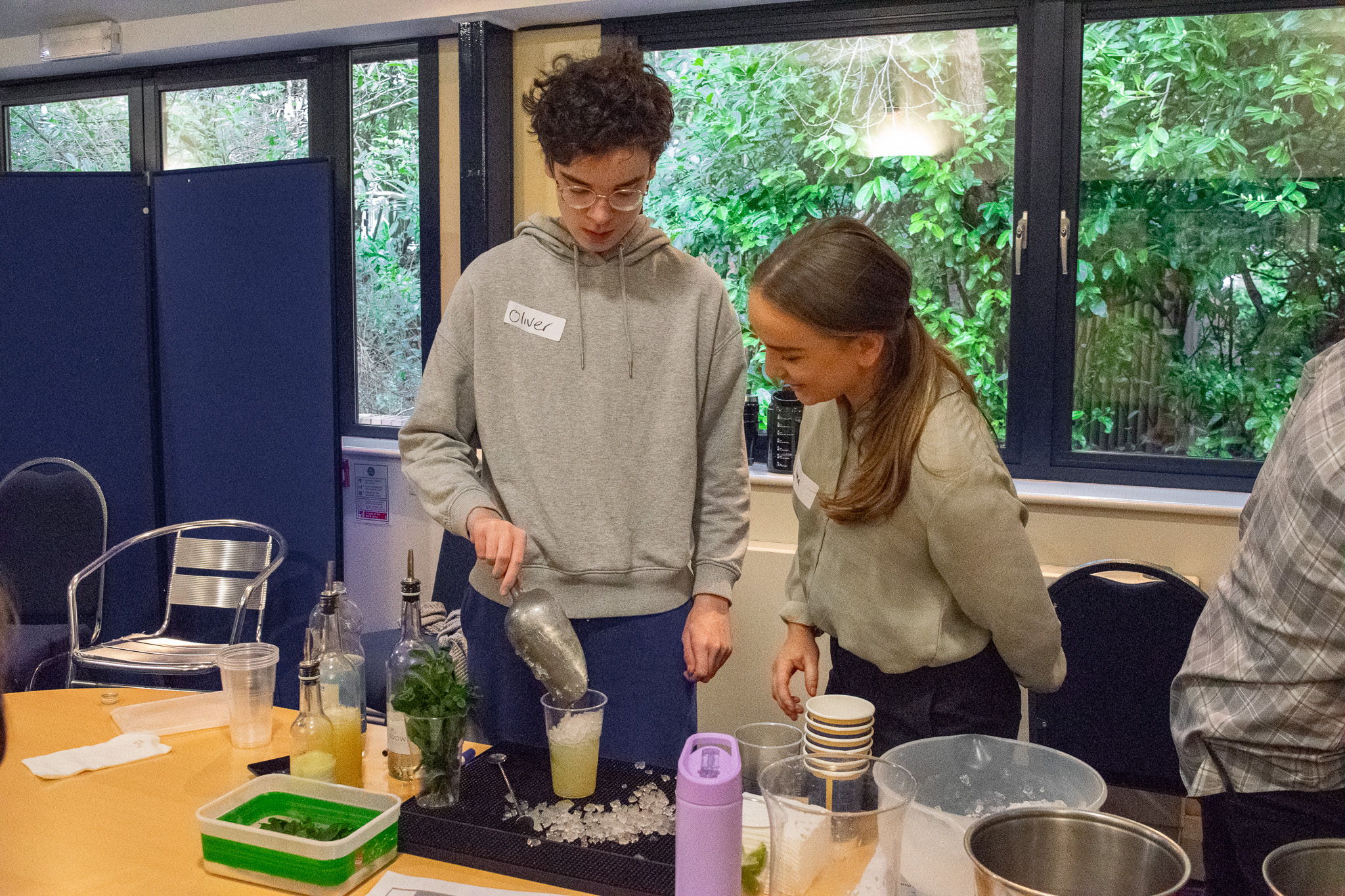 A boy is pouring ice into a cup half-full with alcohol-free mojito. there is a trainer to his right watching and guiding. 