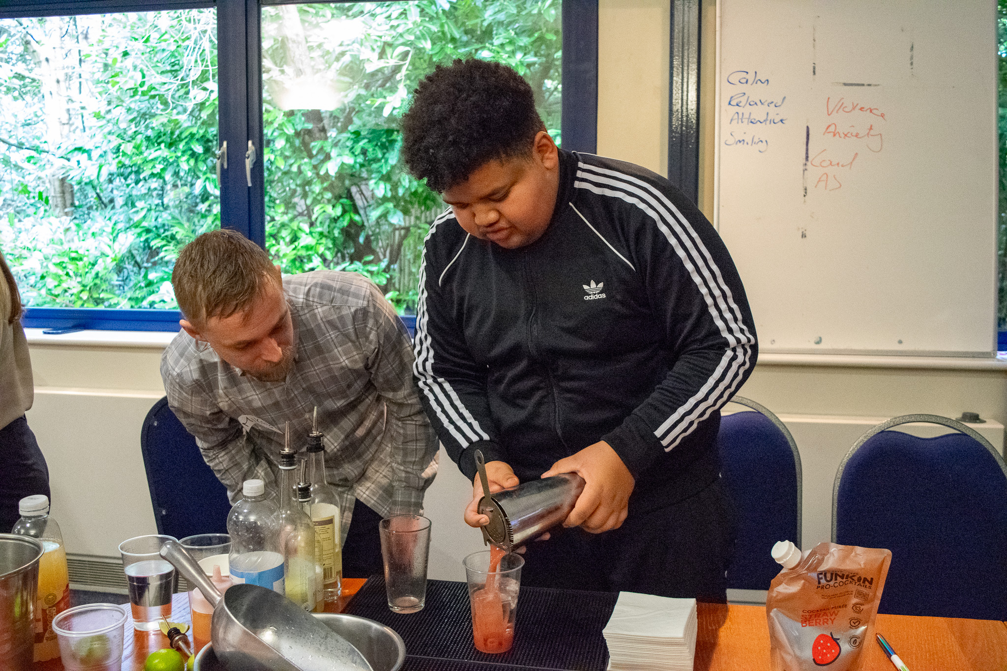 Boy pouring a strawberry soda cocktail from a shaker into a cup. The trainer is standing next to him observing. 