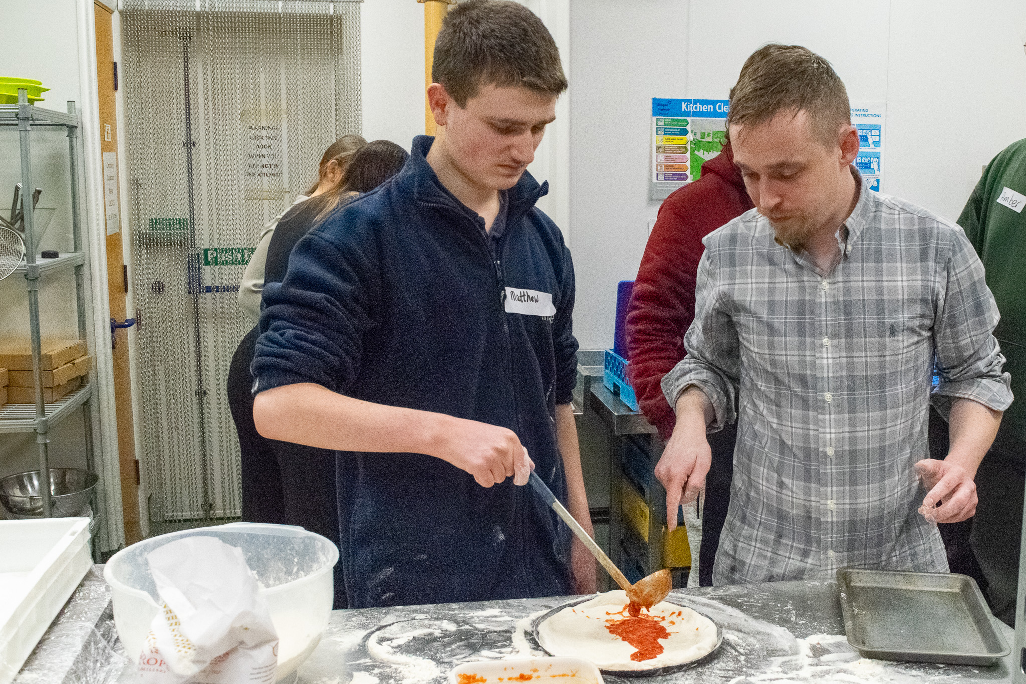 Student holding a ladle of tomato sauce pouring it over his shaped pizza dough on a kitchen work surface. To his right, the teacher is instructing him on technique. 