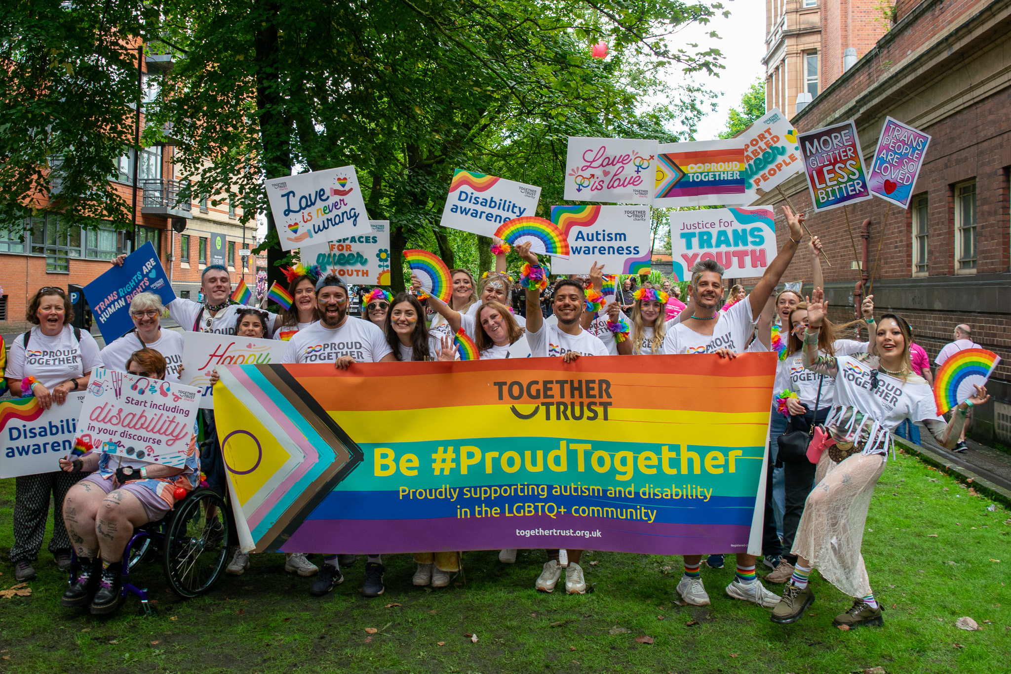 a large Group of people stood in a park holding a rainbow banner that reads "be #proud together. proudly supporting autism and disability in the LGBTQ+ community" 
