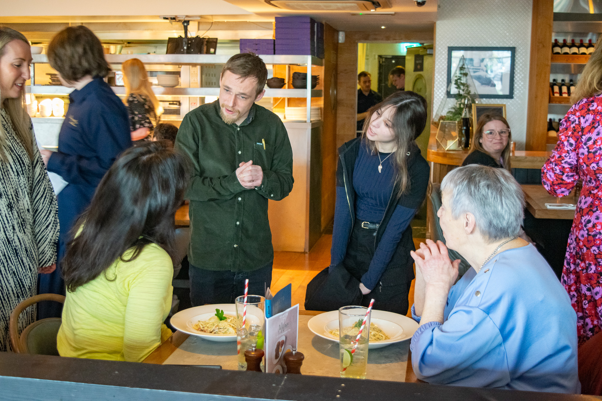 A student stood next to Mikey, Gusto Staff member, talking to a table of customers taking their order. 