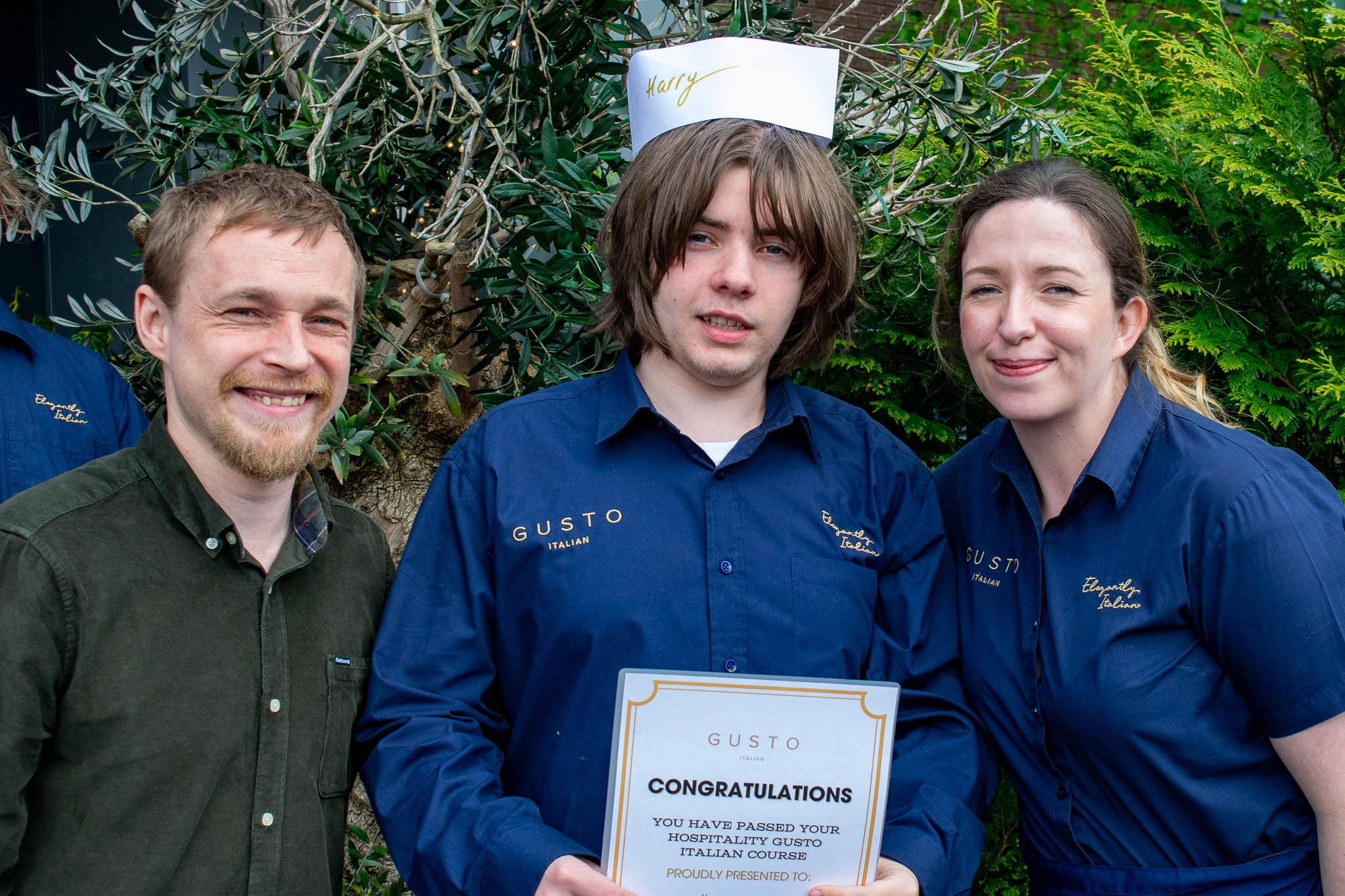 Two staff members from Gusto posing with a student holding a certificate and wearing a paper chef hat.
