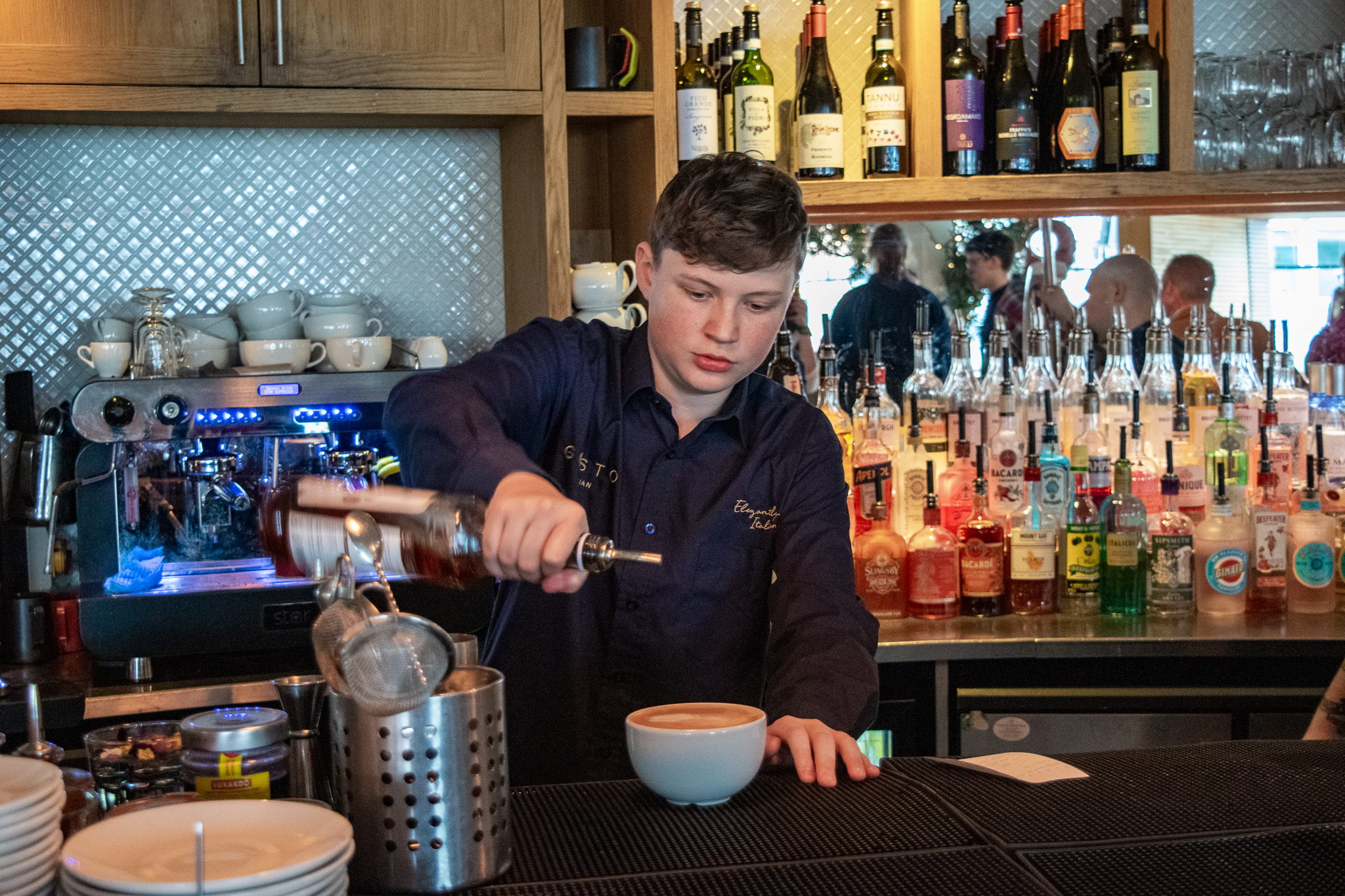 A young person working behind a bar adding syrup to a latte. 