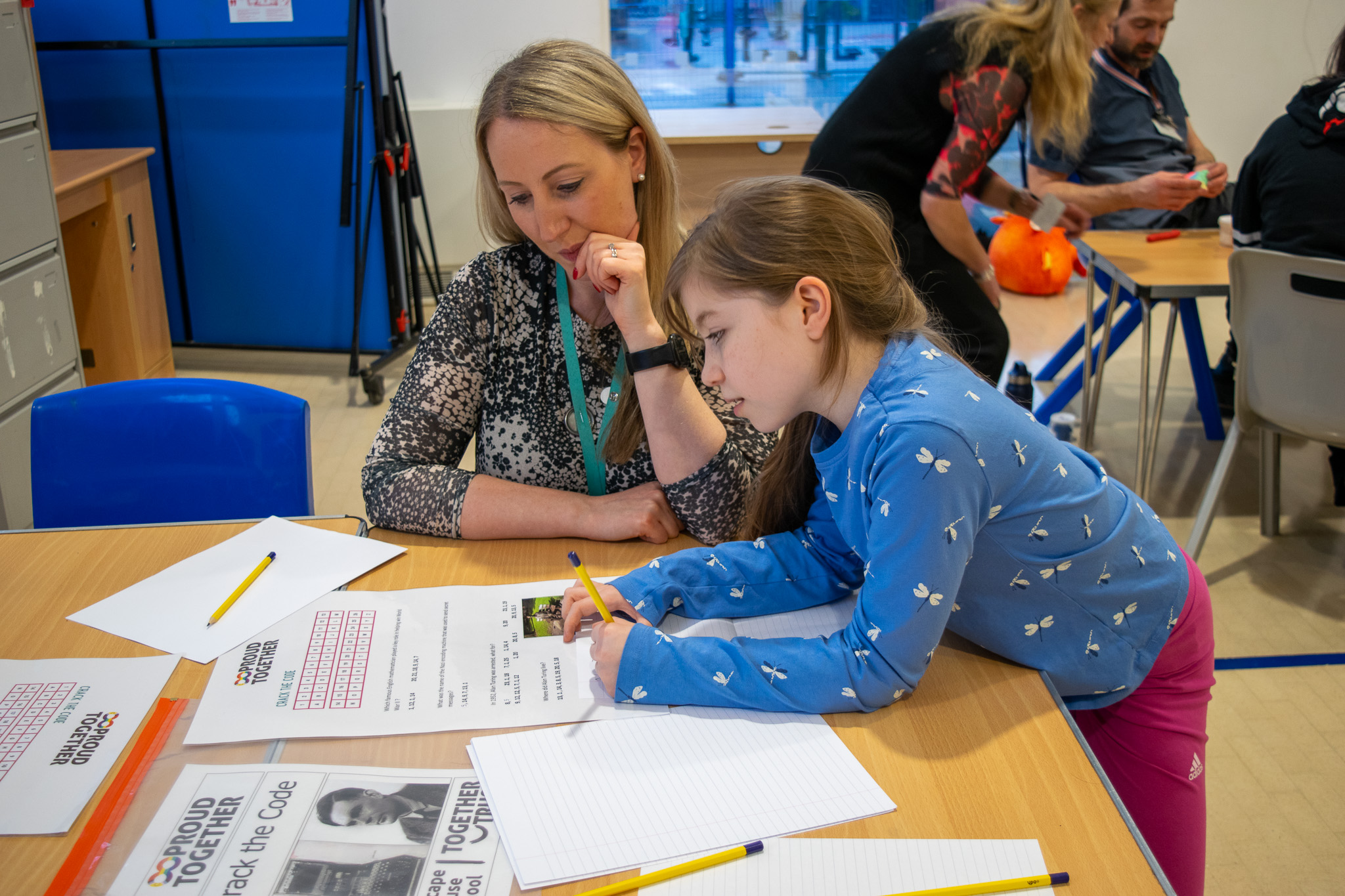 Young girl leaning over a table solving a paper puzzle. Next to her, the school headmaster is also looking at the paper quizzically.