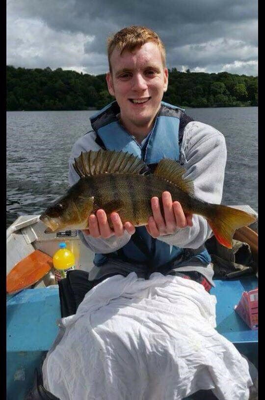 Jordan holding a large fish he has caught while sat in a boat with a large lake visible behind him