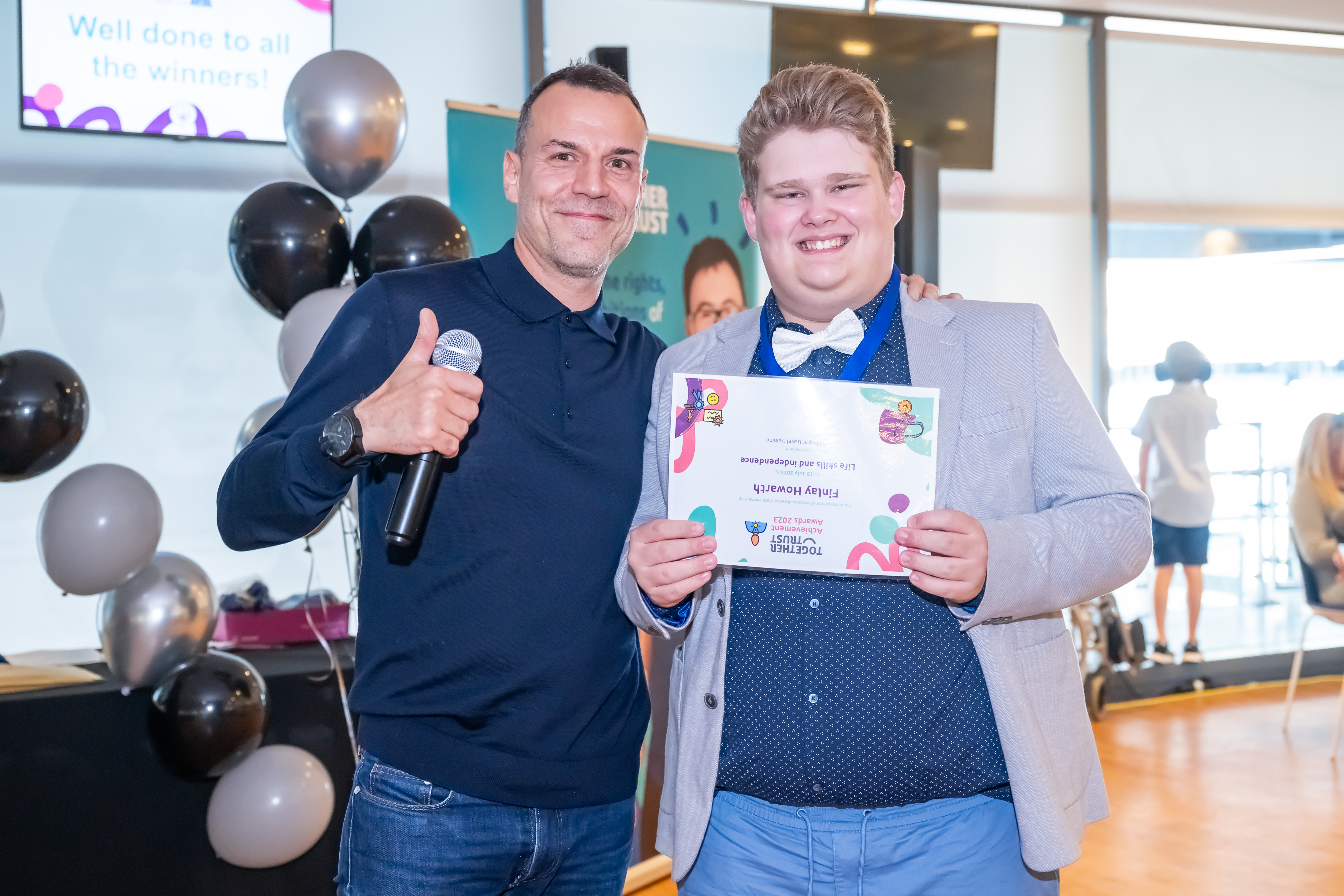 Staff member showing thumbs-up whilst posing next to a young man, holding his certificate