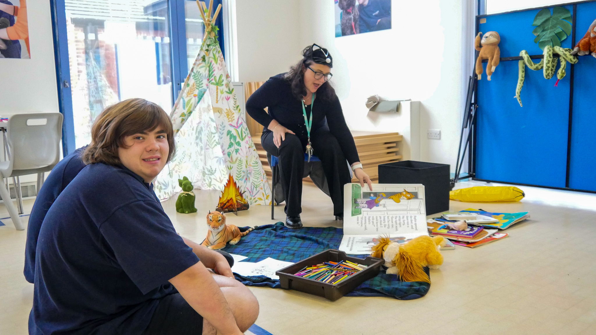 A student sitting down by the colouring and storytelling station. A teacher is reading him a story. 