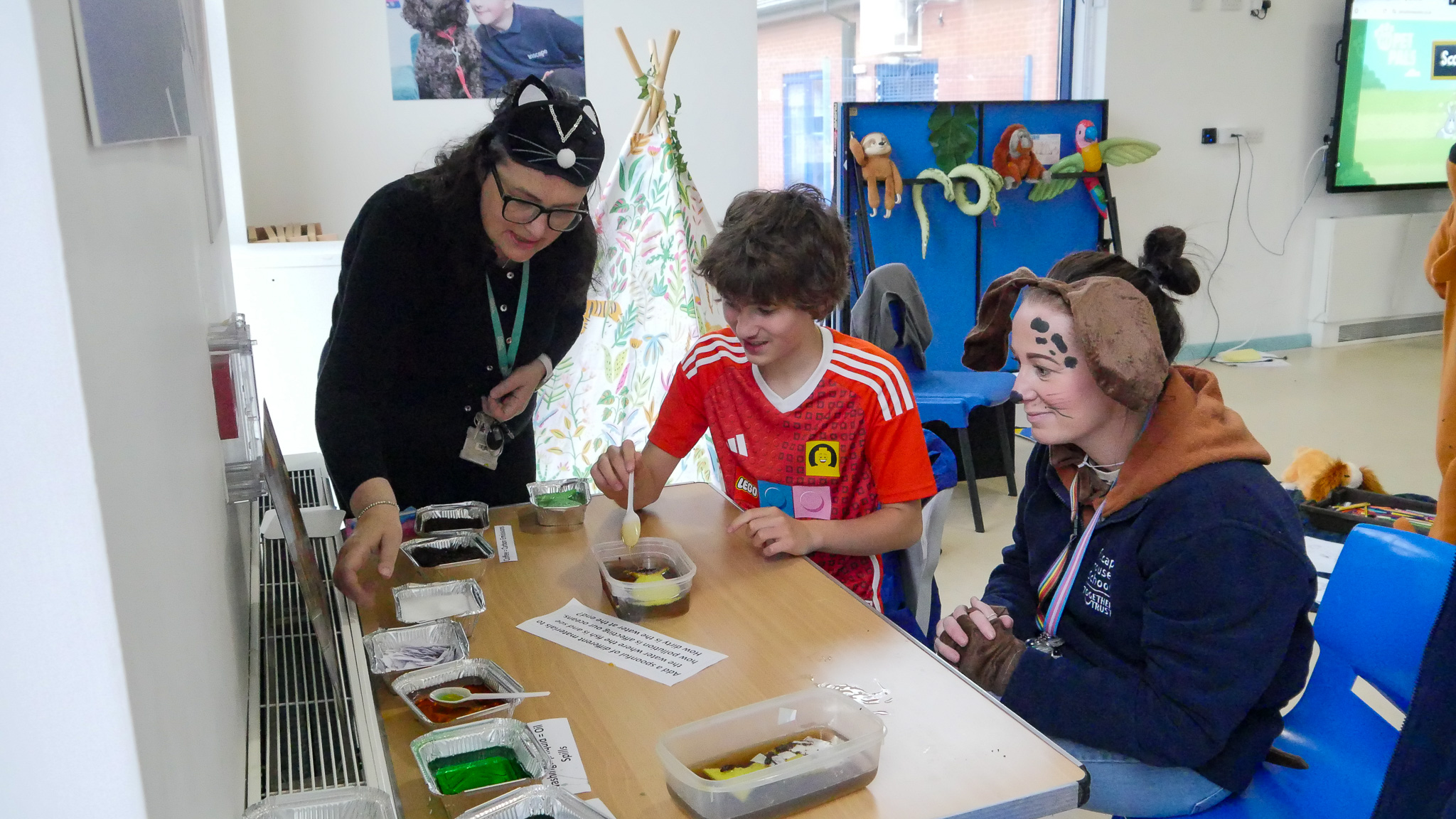 A student interacting with the ocean pollution activity. He is dripping honey into a tub of water. next to him, two teachers are explaining the activity. 