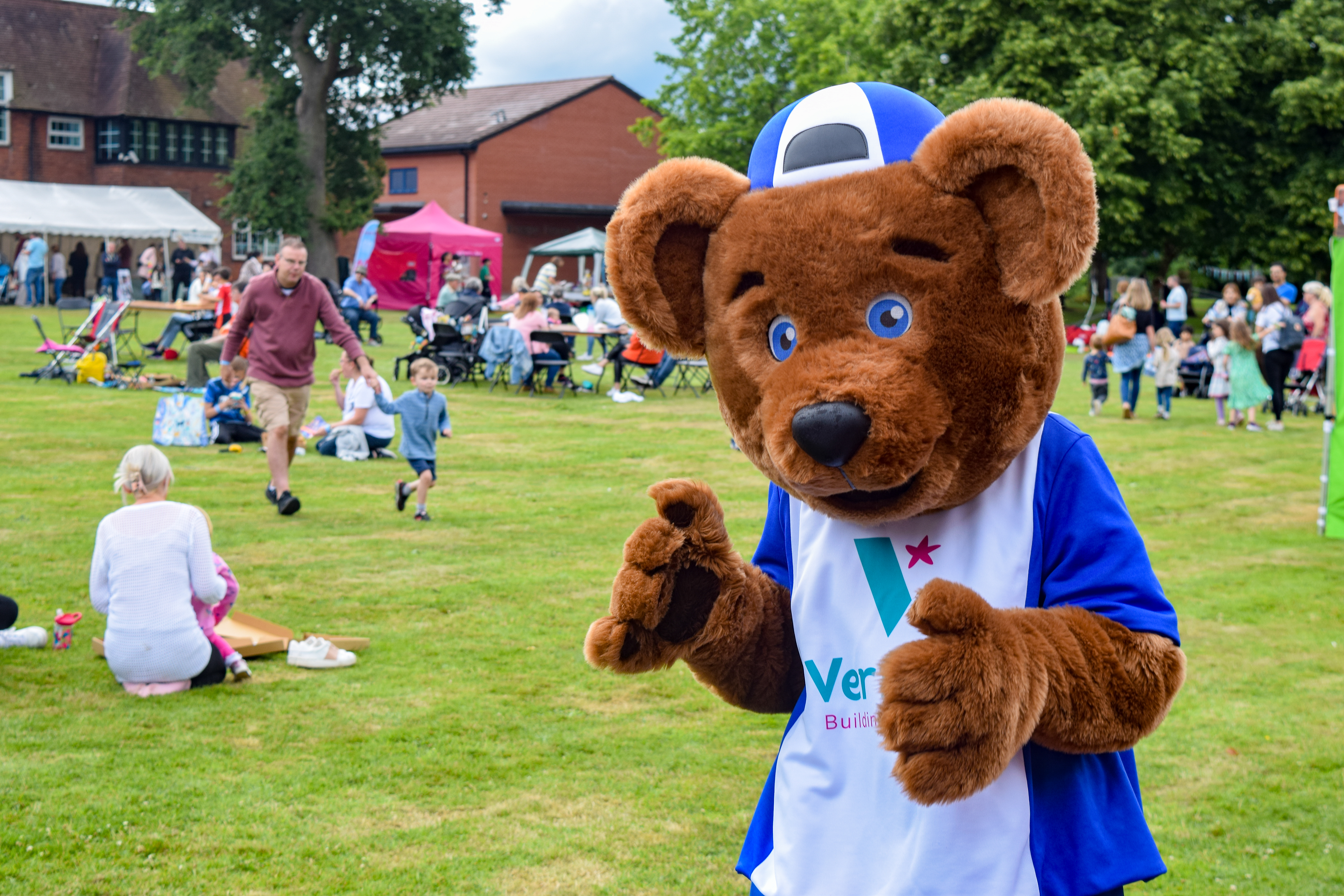 Teddy bear mascot waving at the camera in front of a lawn full of people at a festival. 