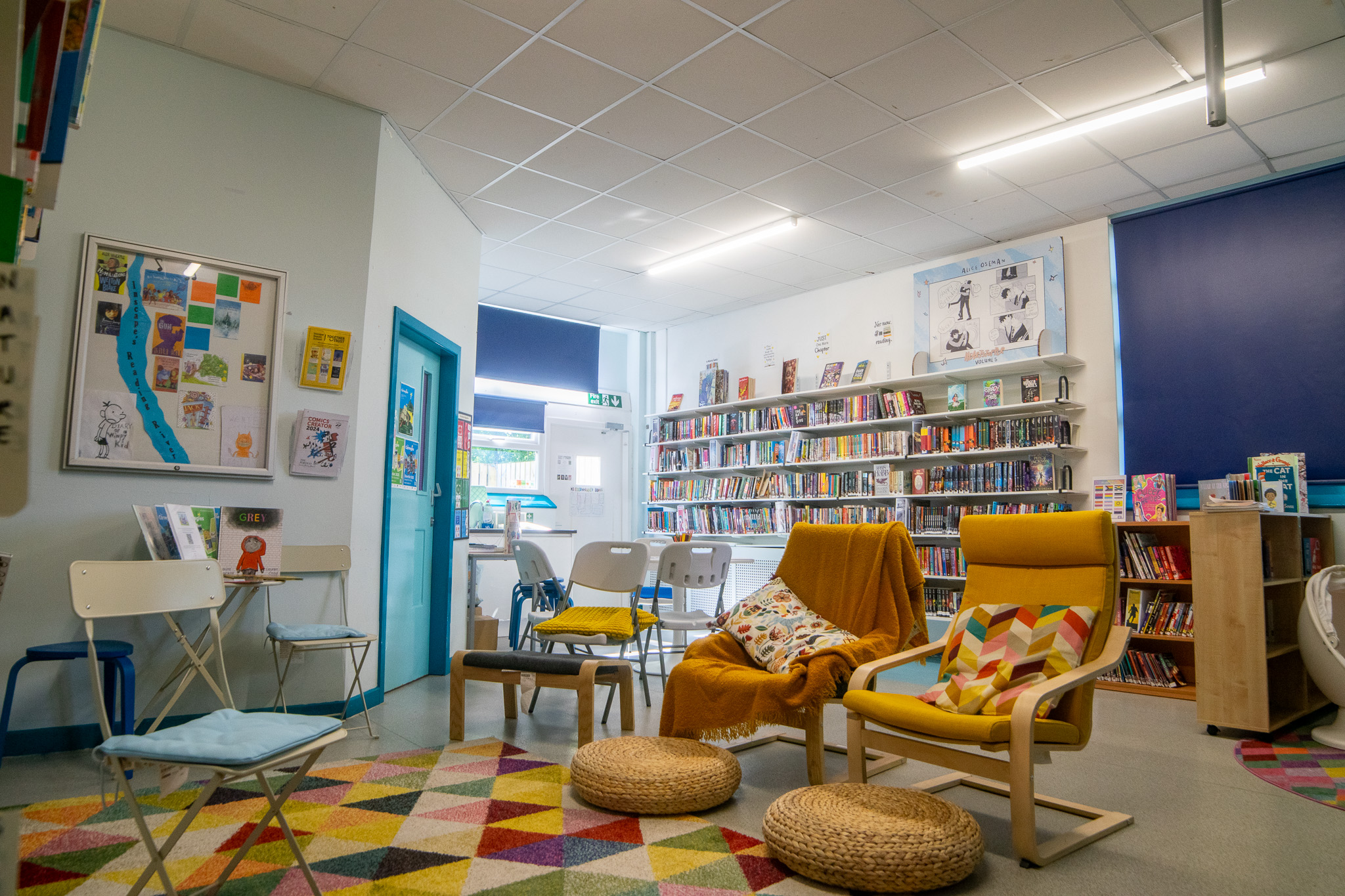 A wide view of the library that has 2 big armchairs, a colorful rug, cute cushions, and walls full of books. 