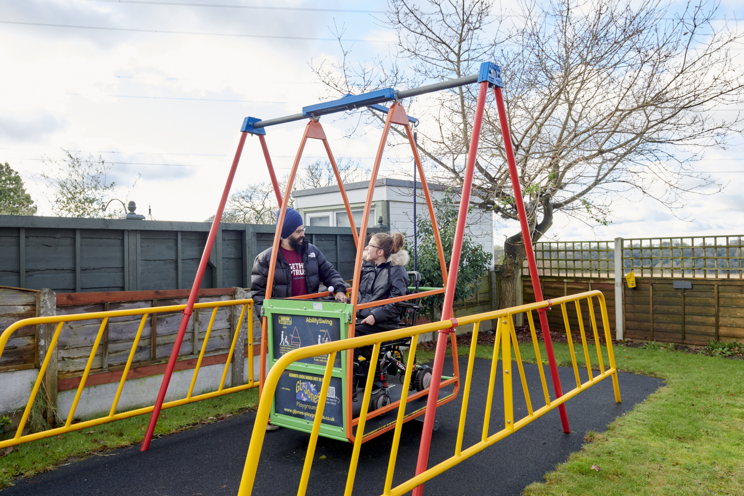 A young girl in a wheelchair on an accessible swing, being helped by her support worker.