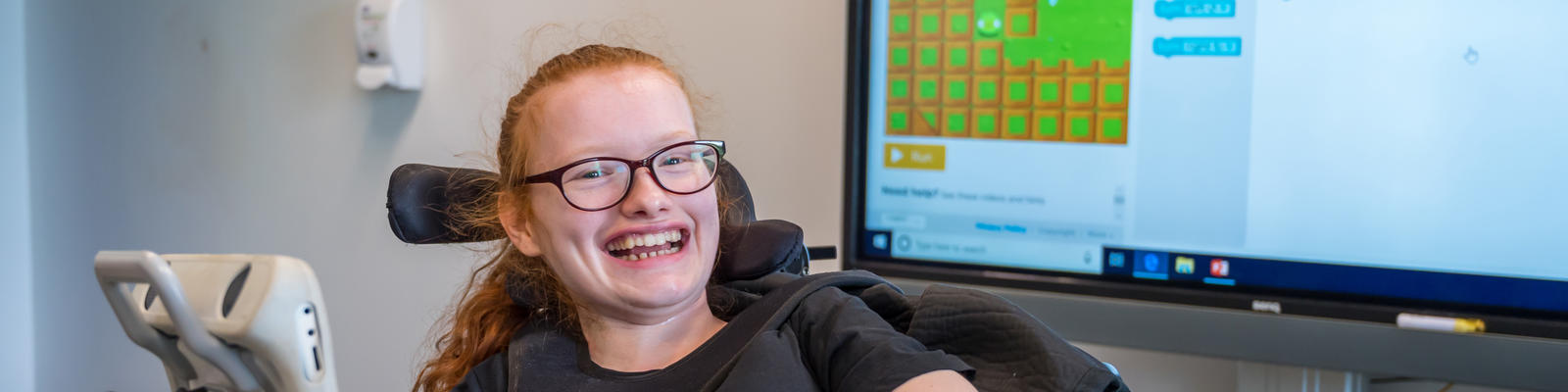 Young girl in wheelchair smiling wide at the camera. She is sat in front of a big interactive screen in a classroom. 