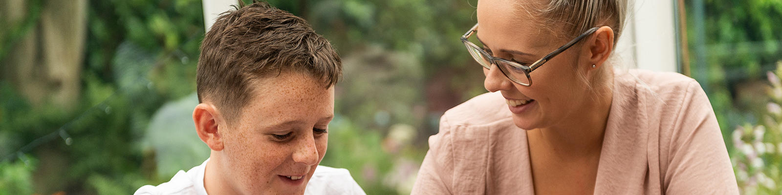 Woman smiling at a young boy. They are both sat at a table in a house