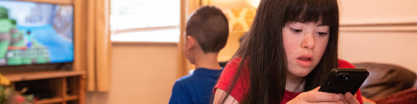 Young girl sitting down on couch holding a mobile phone. Young boy behind her watching the TV 