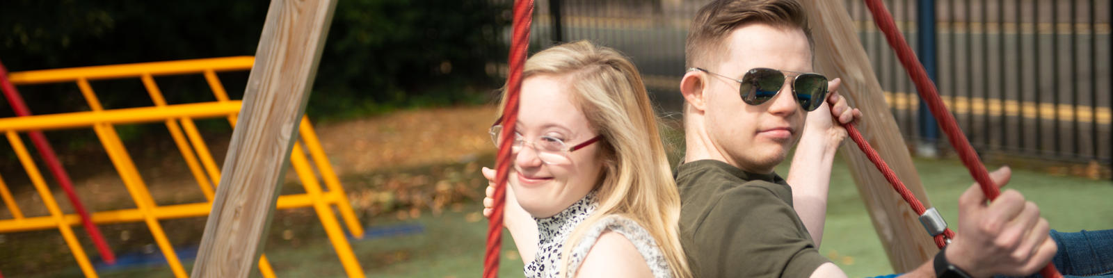 Two young people with down syndrome, a girl and a boy sat back to back on a swing.