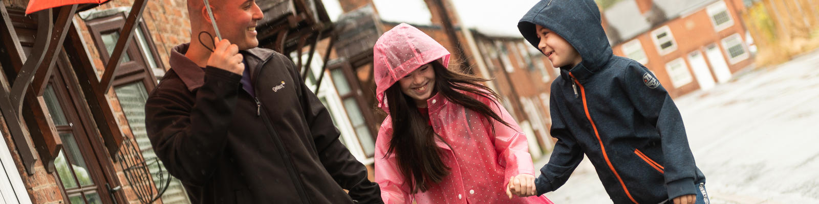 Adult man walking two young people through the rain as he is holding an umbrella over them. They are all smiling.