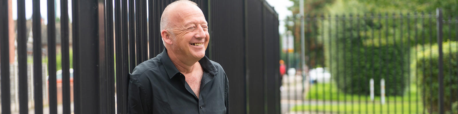 Man smiles looking in the distance. He is standing outside leaning on a fence 