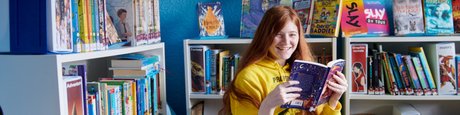 Young girl with long ginger hair sitting down on a chair in the library reading a book.