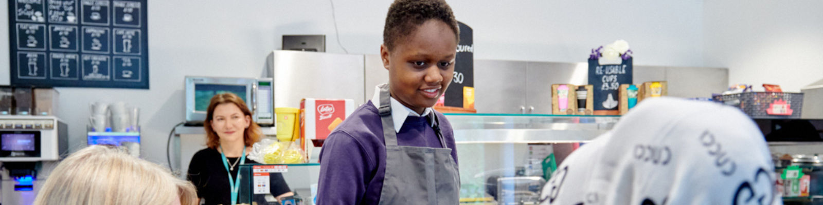 A young person serving drinks in the school cafe to 2 women sat down. 