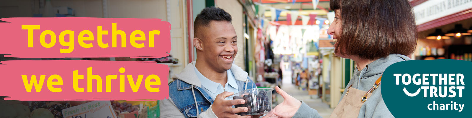 Young man with down syndrome smiling at a shop keeper. She is handing him a punnet of berries. They are in an indoor market. 