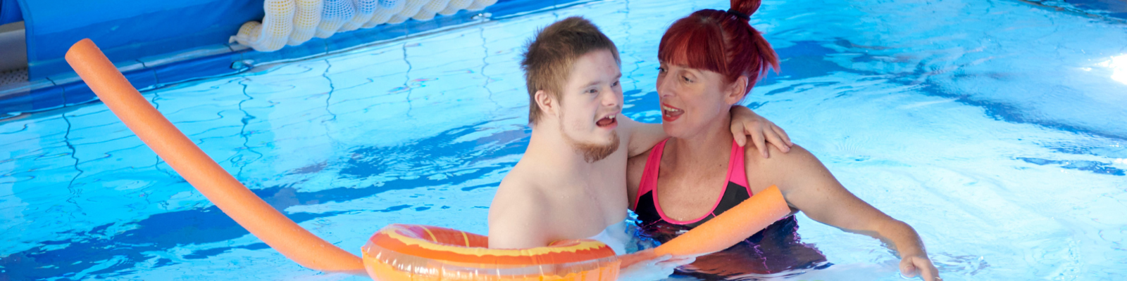 Man with disability floating in a pool assisted by social carer and pool noodles. They are both smiling wide.