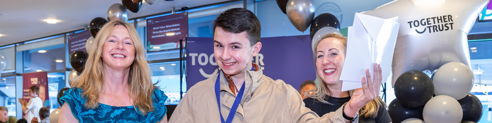 Student wearing a medal smiling wide holding a diploma up. Next to him are two teachers smiling at him. 