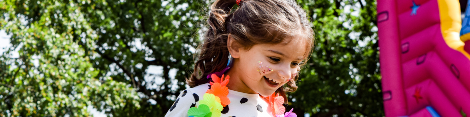 Young girl wearing a flower crown around her neck smiling. She is outside in a park. 