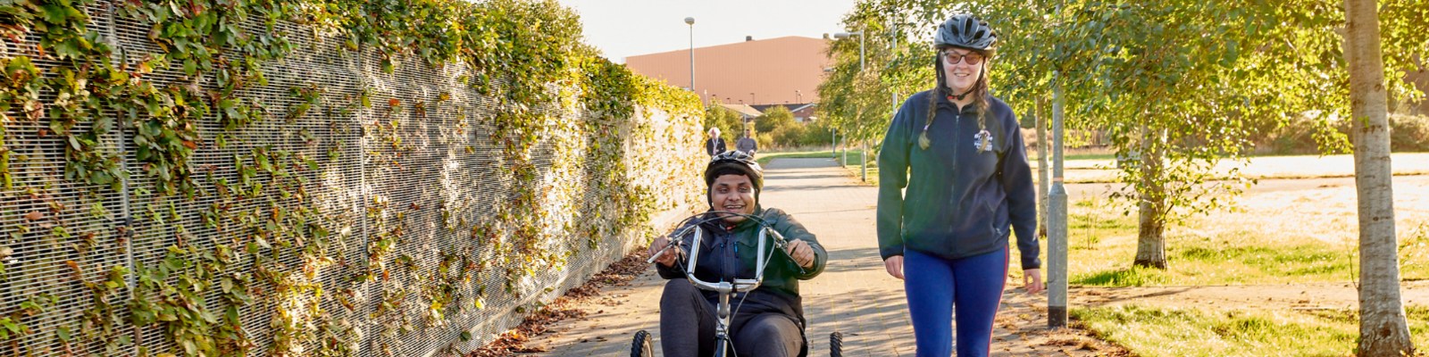 Man cycling in an adaptive bike next to his support carer in Bridge College courtyard.