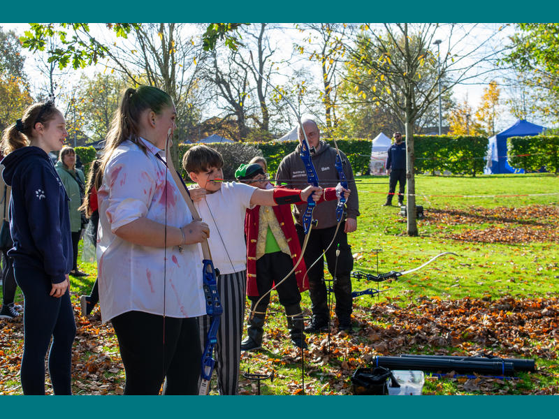 A row of young people in fancy dress trying their hand at archery against a sunny backdrop.