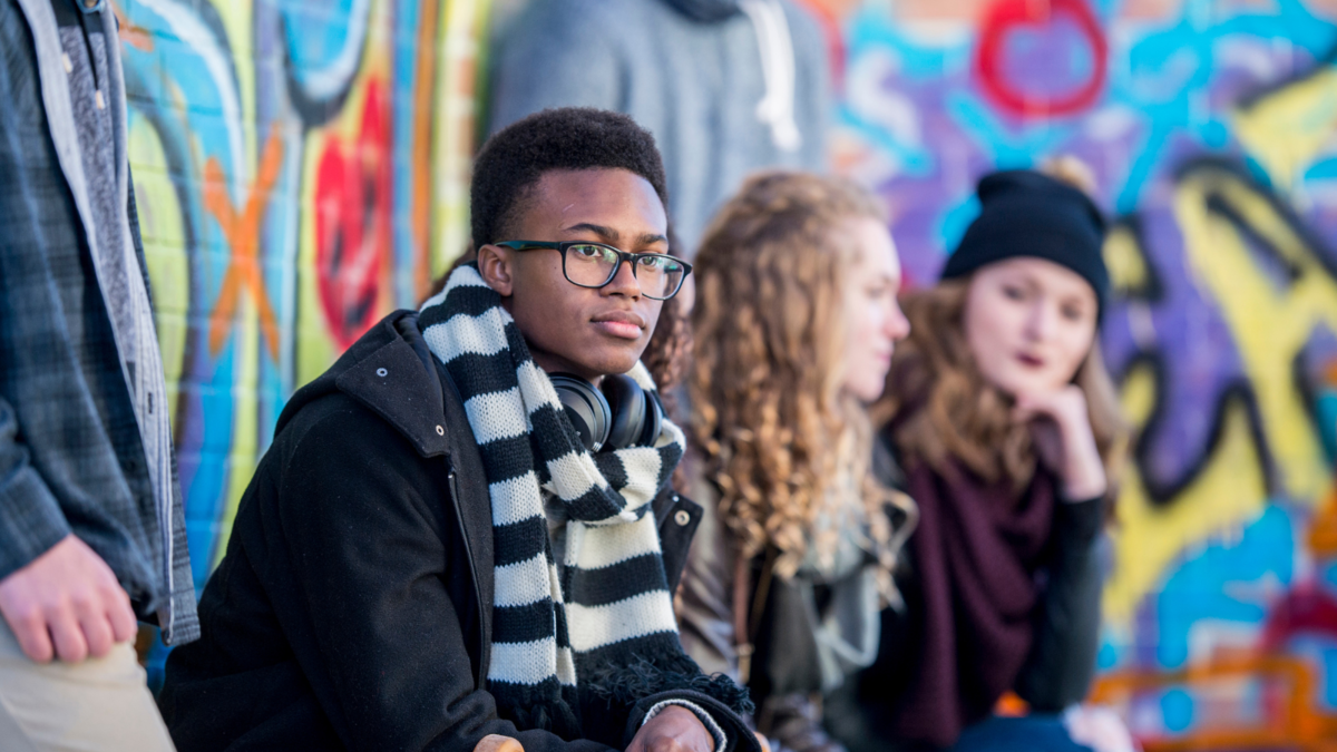 Young man looking pensive, sat next to two others in front of a wall painted with bright graffiti.