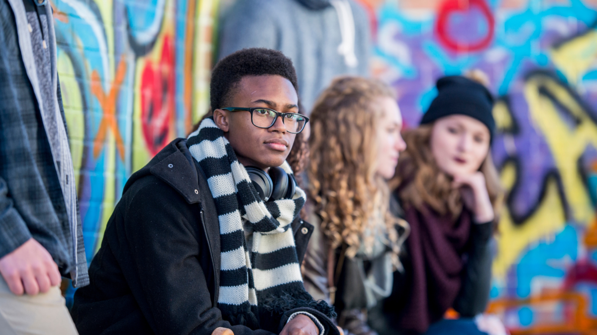 Young man looking pensive, sat next to two others in front of a wall painted with bright graffiti. 