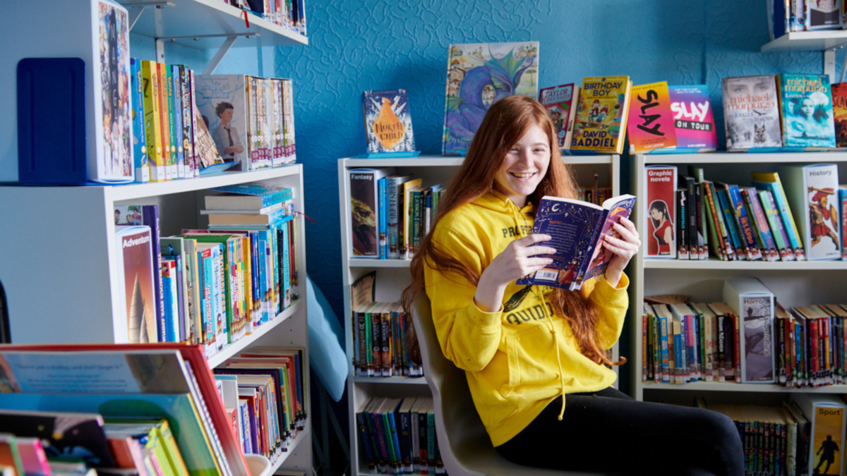 A teenage girl holding up a book smiling in the school library