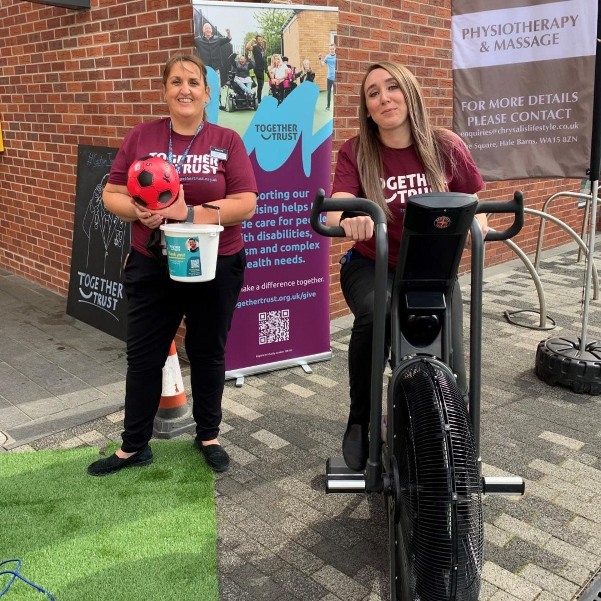 A team member from Booths holding a collection bucket next to another sat on an exercise bike A team member from Booths holding a collection bucket next to another sat on an exercise bike