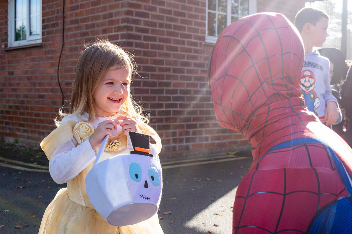 Young girl dressed as a Princess carrying a trick-or-treat bucket, talking to Spider Man.