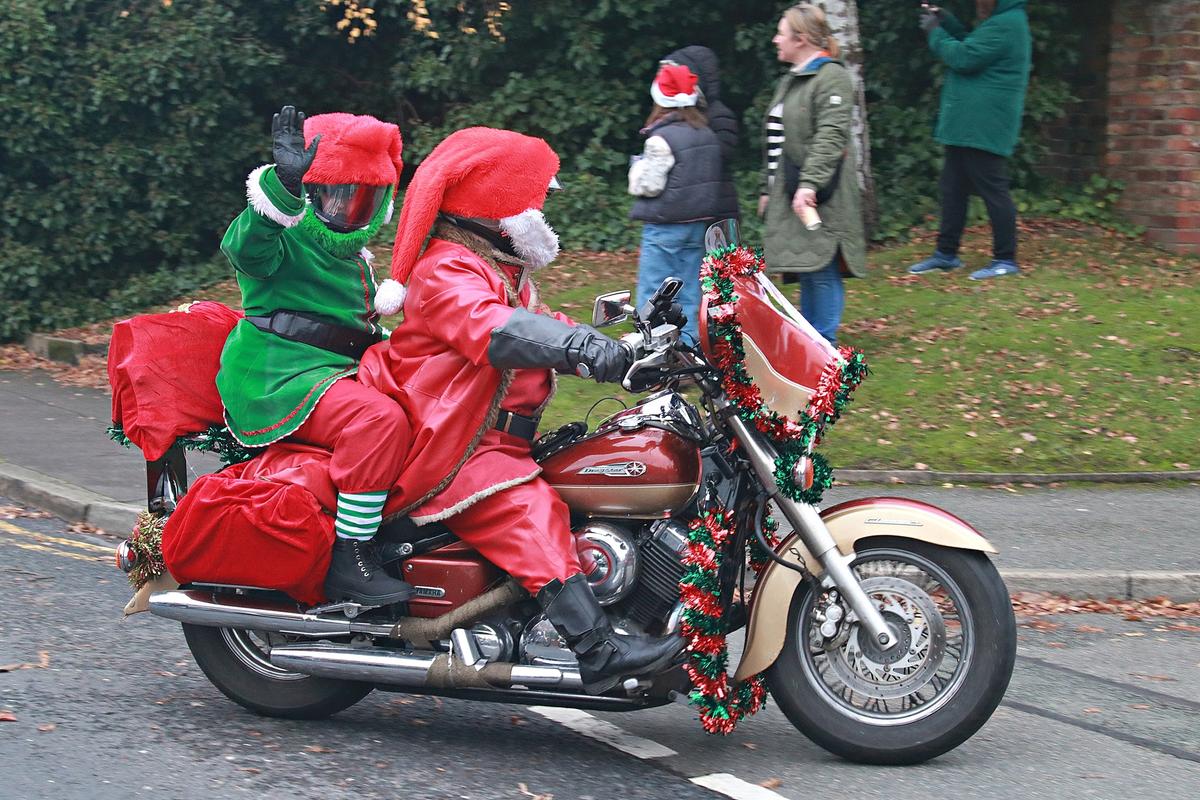 Two bikers on a motorcycle, dressed up as Santa Claus and an elf, waving cheerfully at the camera