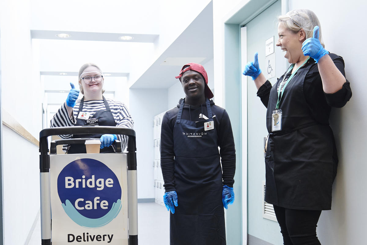 two bridge students pushing a cart with drinks smiling with a member of staff