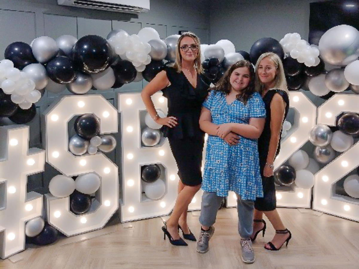 Alice wearing a blue dress standing in front of a light up sign alongside her 2 teachers wearing black dresses. They are all smiling wide. 