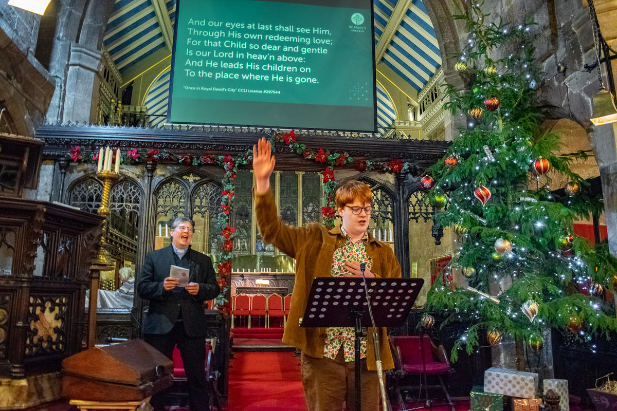 Teenage boy wearing a corduroy blazer singing. He is holding one hand up. He is stood in front of a music stand in an old church. Behind him is a screen with lyrics to the song. 