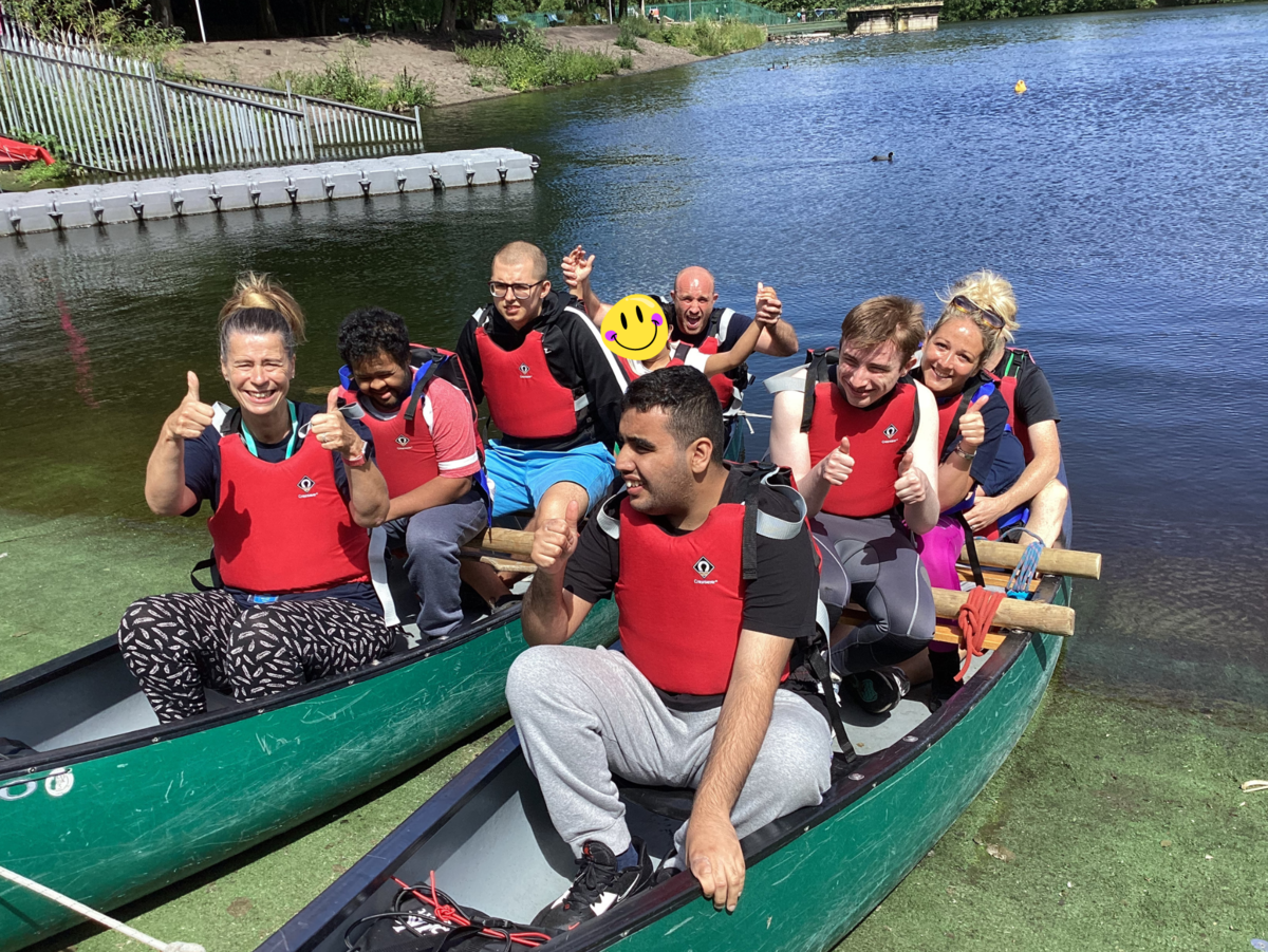 Two kayaks on the edge of the water full of students and staff wearing red life jackets, smiling and waving at the camera