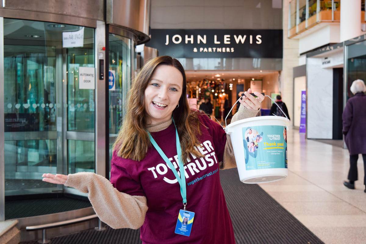 Woman wearing a purple together trust tshirt standing inside a John lewis store. She is holding a donation bucket and smiling wide. 