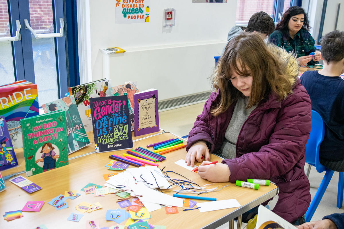 Young girl drawing on a bookmark at a table full of books and stickers. 