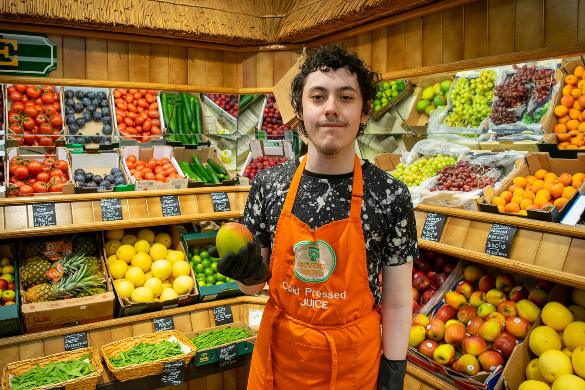 A teen boy wearing an apron is smiling in front of a well-lit wall of fresh fruit. He is holding a mango and smiling. 
