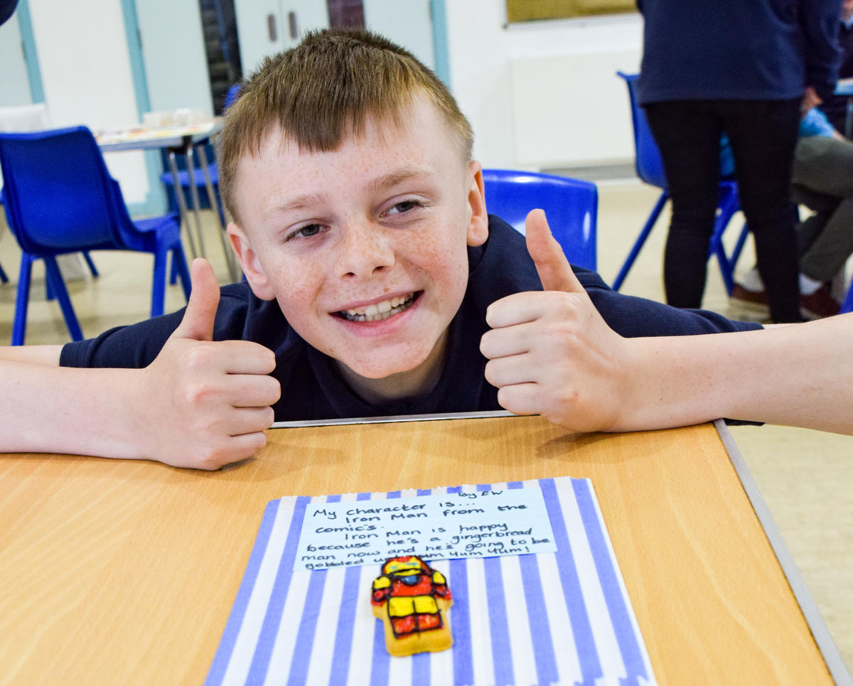 young boy smiling wide with both of his thumbs up. he is standing above a gingerbread man decorated to look like Iron Man