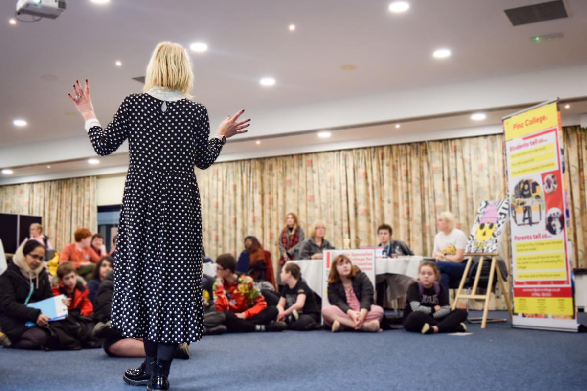 Woman wearing long dress standing in front of a group of children. She is talking to them pointing to a screen. The children are all sat on the floor looking up. 