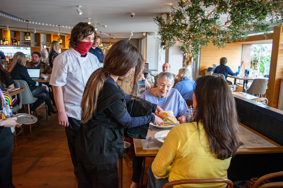 Two young people presenting a portion of pasta to a table of customers. One of them is grating cheese over the pasta. 