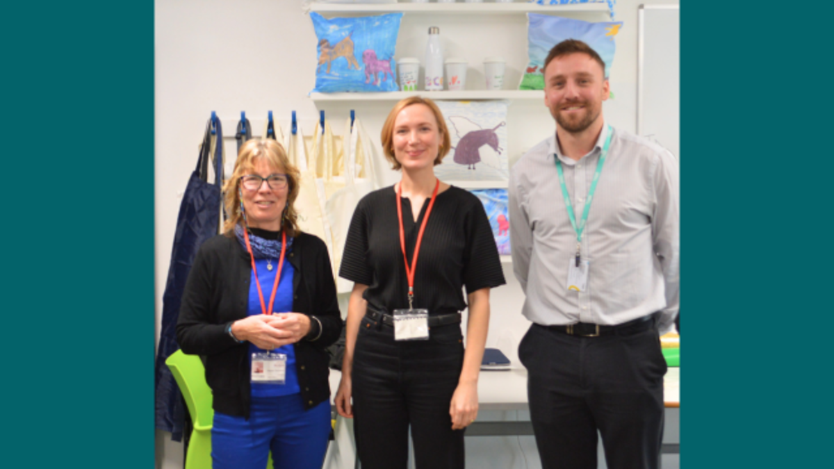 A photograph showing Maggie, Kate, Jeremy stood in a class room in front of shelves displaying students print work including, cushions and water bottles